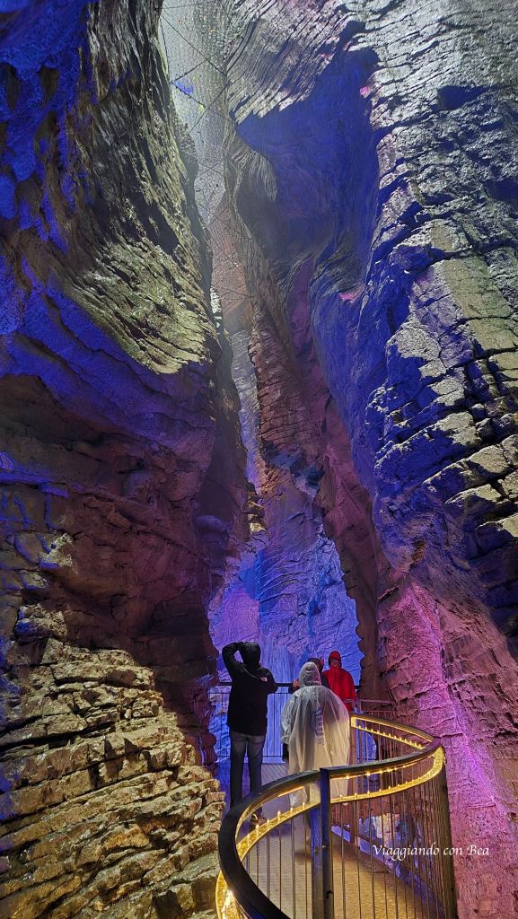 Interno di una gola con pareti di roccia illuminate da luci colorate, persone che esplorano il sentiero con parapetto.