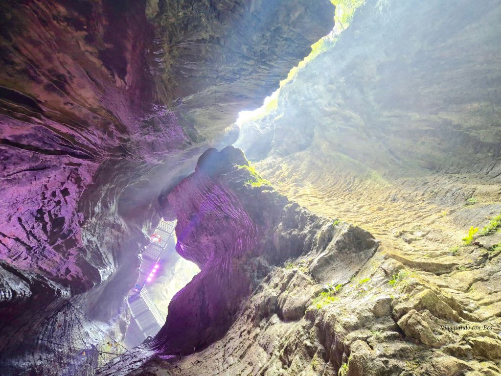 Vista dell'interno di una grotta con pareti rocciose illuminate da luci viola, che porta verso l'uscita in alto.