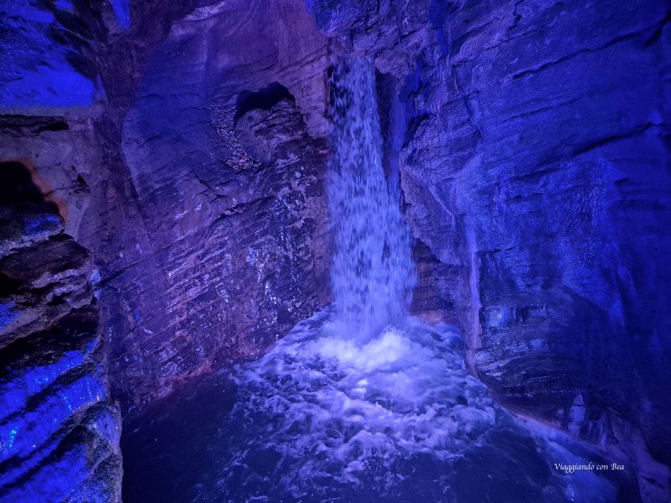 Cascata che scorre in una grotta illuminata da luci blu, con pareti di pietra bagnate.